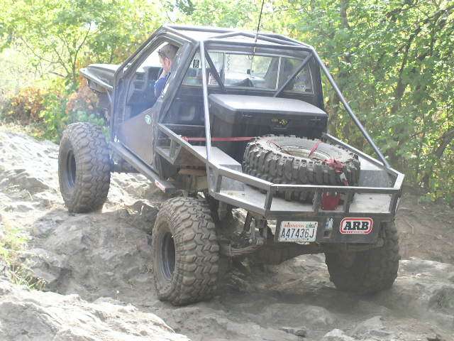 Doorless caged gray 4x4 with bed-mounted spare tire crawling a rock ledge