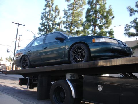 1995 Ford Taurus - A dark green sedan with aftermarket wheels is being transported on a tow truck.