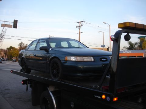 1995 Ford Taurus - A dark green Ford Taurus is loaded onto a tow truck, seen from the front-right.