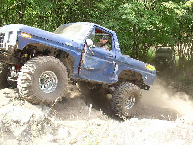 Blue topless full-size Bronco on huge mud tires climbing a dirt bank in the woods