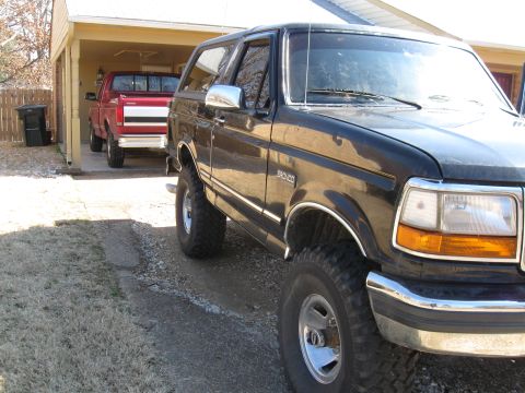 1994 Ford Bronco - A black Ford Bronco is parked in a driveway next to a red pickup truck under a carport.