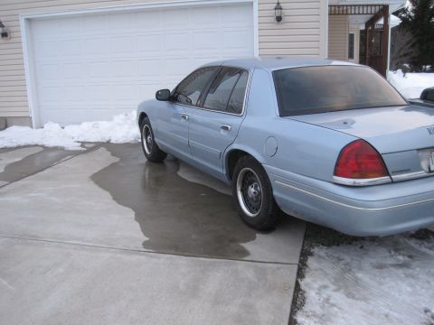 1999 Ford Crown Victoria - A light blue sedan is parked on a wet driveway in front of a garage with snow on the ground.