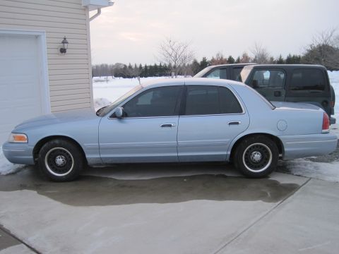 1999 Ford Crown Victoria - Side view of a light blue 1999 Ford Crown Victoria sedan parked on a concrete driveway next to a garage, with another vehicle visible behin…