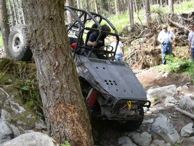Off-road buggy tilted against a tree on a rocky trail with people watching