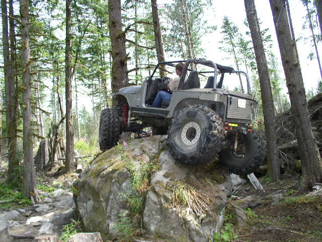 Caged doorless tan Jeep on huge mud tires flexing over a rock ledge in the woods