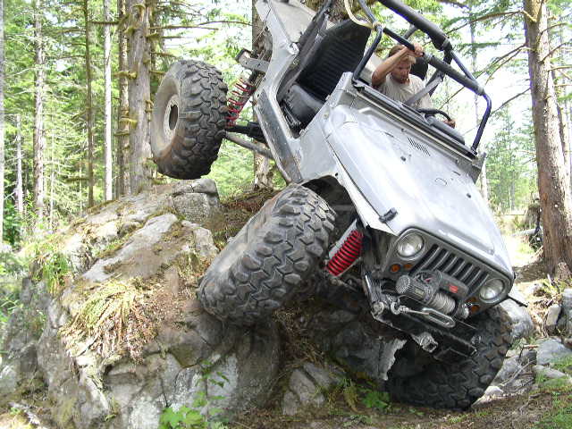 Silver Jeep Wrangler with front winch and red shocks flexing over a mossy rock ledge