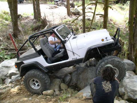 1979 Ford Bronco - A white Jeep Wrangler is shown off-roading over large rocks, with a person watching from the side.