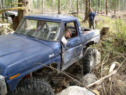 1979 Ford Bronco - A blue pickup truck with oversized tires is shown off-roading through a wooded area, with another vehicle in the background.