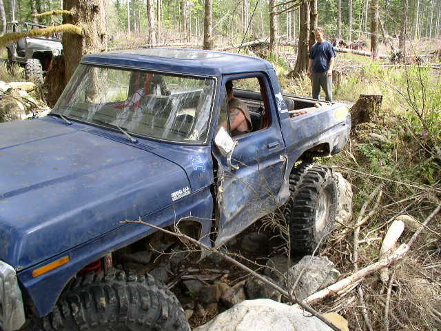 Blue topless lifted Ford Bronco flexing over rocks on a wooded trail