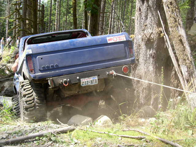 Blue lifted Ford pickup on mud tires winching off a rocky forest trail, rear view