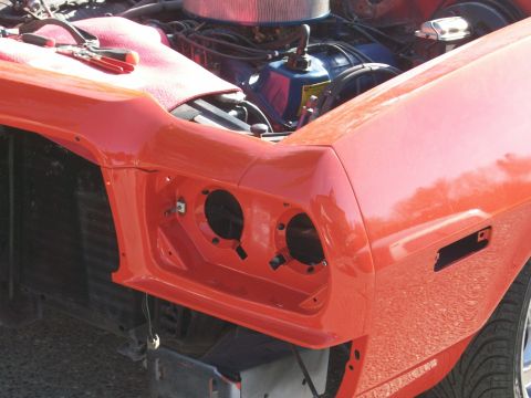 1973 Ford Ranchero - Close-up of the left front corner of an orange-red car mid-assembly, with the dual headlight buckets empty, the grille removed and the radi…