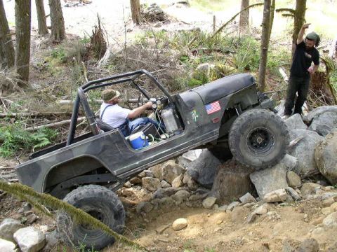 1979 Ford Bronco - A black Jeep CJ-7 navigates a rocky trail with a driver behind the wheel and a person standing on the rocks nearby.