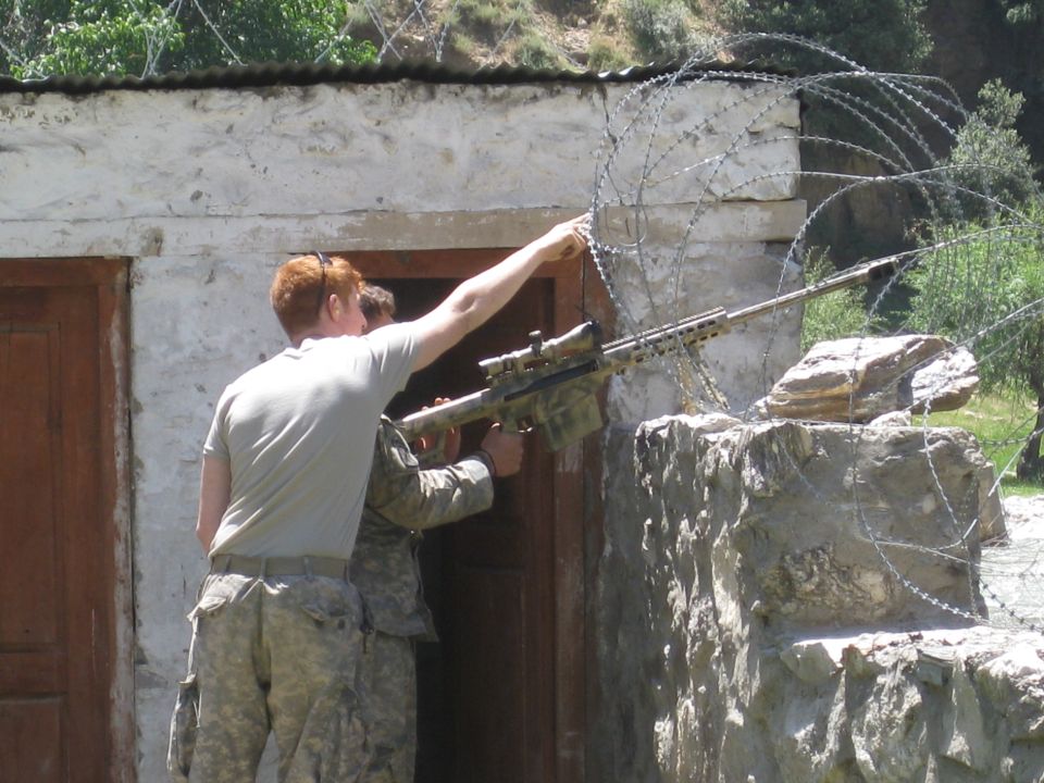 Soldiers aim sniper rifle over barbed wire wall near stone building