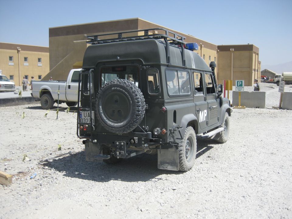 Dark green Land Rover Defender with MP markings, spare tire on back, parked near buildings. White pickup truck in background