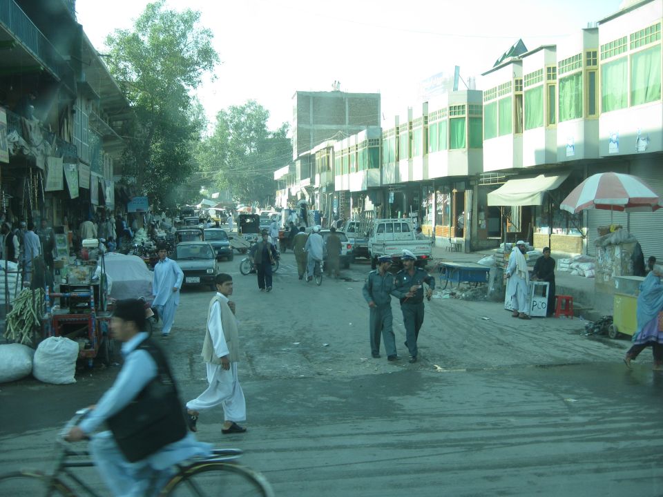 Street scene in a market with people walking, riding bicycles, and several cars and trucks parked or driving