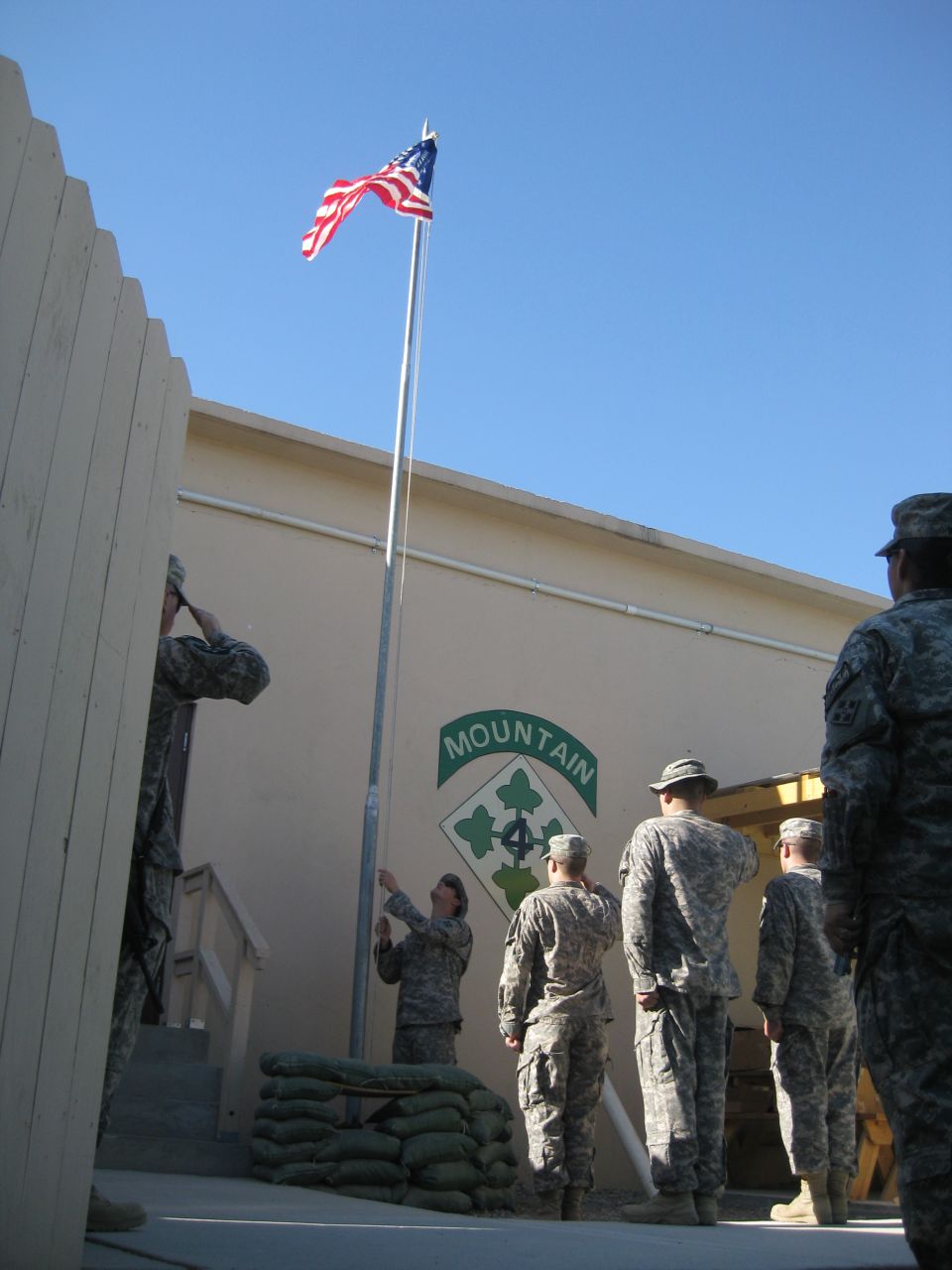 Military personnel salute the American flag being raised on a flagpole in front of a building with a unit insignia