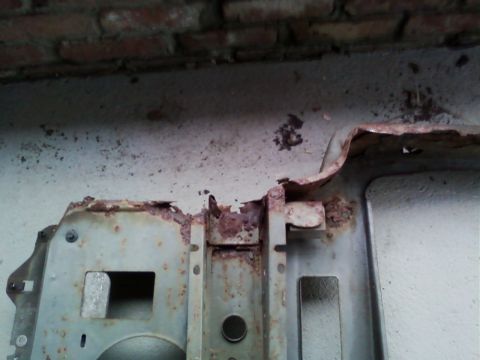 1990 Ford Bronco - Close-up of a rusted sheet-metal radiator core support panel laid on a white textured floor against a brick wall, showing heavy corrosion,…
