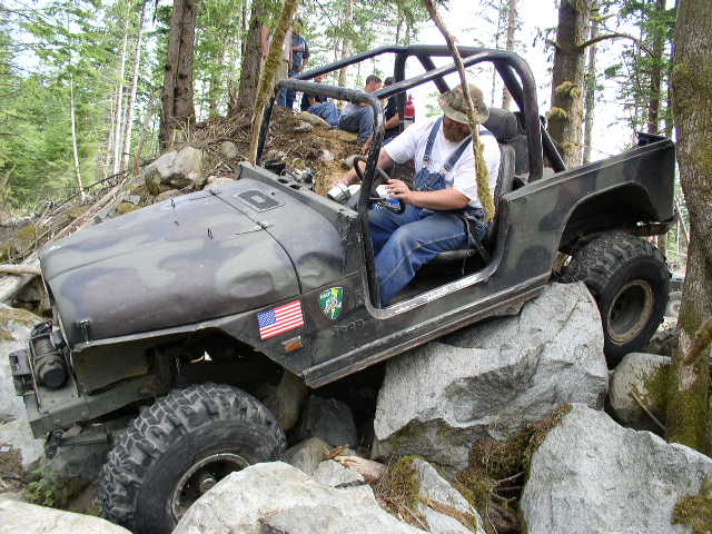 Flat-black doorless caged 4x4 crawling over boulders on a wooded rock trail