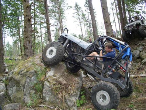 1979 Ford Bronco - An off-road vehicle navigates a rocky incline in a wooded area, with another vehicle visible in the background.