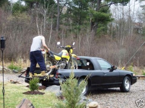 1972 Ford Ranchero - A man loads a yellow dirt bike into the bed of a black sedan pickup truck.