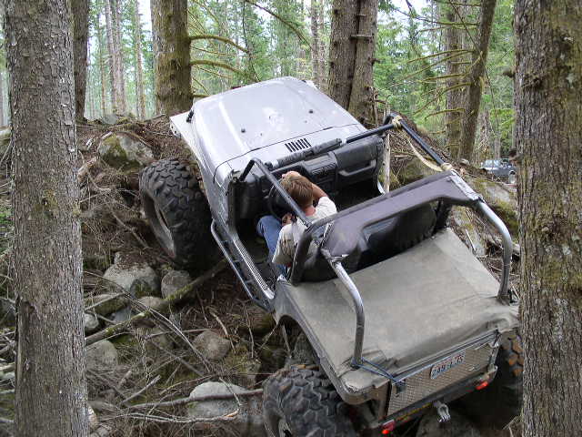 Off-road vehicle climbing over rocks and logs in a forest. Another vehicle is visible in the background