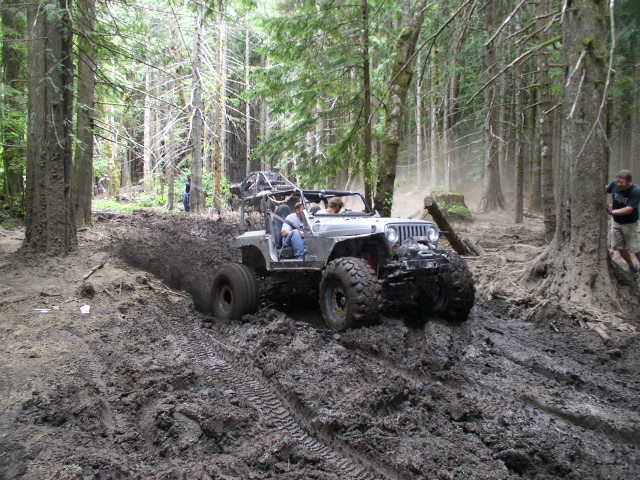Silver caged Jeep CJ on mud tires powering through a deep mud pit in the woods
