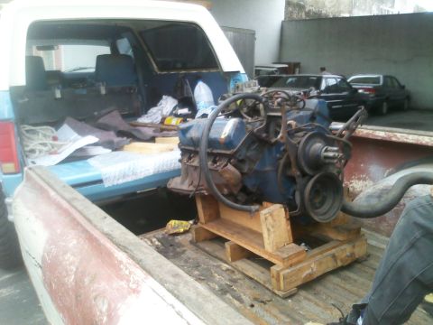 1986 Ford Bronco - An engine is loaded into the bed of a pickup truck, with other vehicles visible in the background.