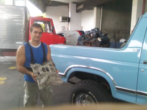 1986 Ford Bronco - A man holds an engine intake manifold next to a light blue 1986 Ford Bronco, with a red truck and engine in the background.