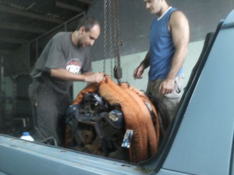 1986 Ford Bronco - Two men are working to load an engine into the back of a vehicle using a chain hoist.