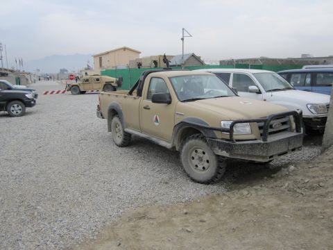 2003 Jeep Wrangler - A tan pickup truck with a black roll bar and bull bar is parked in a gravel lot, with other vehicles and buildings in the background.