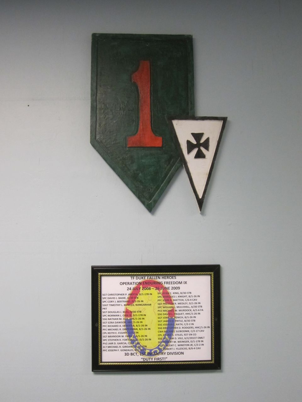 Green shield with red 1, white triangle with black cross, and framed memorial listing names and dates