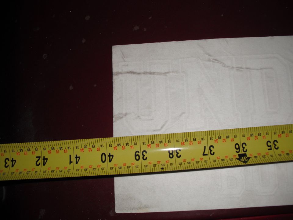 White sticker with faint outline of letters, measured with a yellow tape measure showing inches from 35 to 43