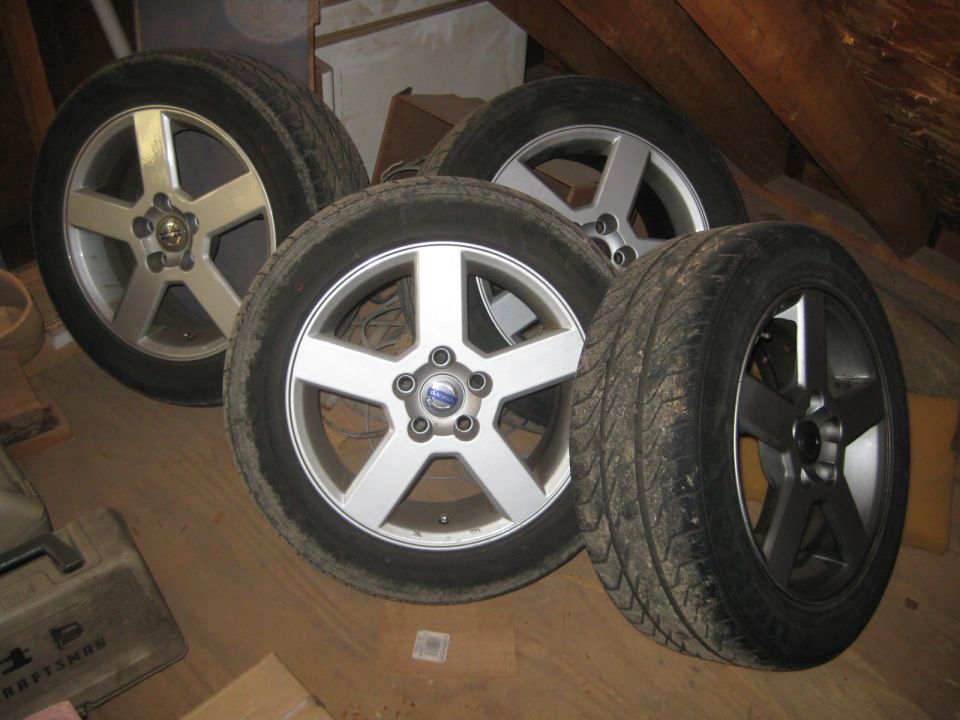 Four tires and wheels, three silver with five spokes and one dark gray with five spokes, on a wooden floor