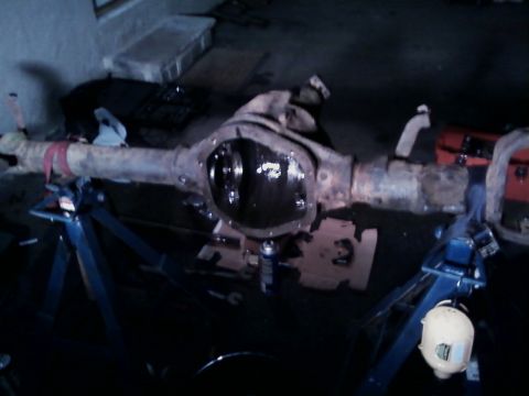 1990 Ford Bronco - A rust-covered axle housing set up on blue jack stands in a dim garage with the differential cover off, showing the cleaned gasket flange a…