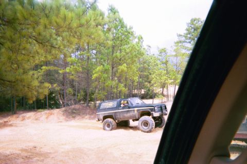 1985 Ford Bronco - A lifted black SUV with large tires is parked on a dirt road surrounded by trees.