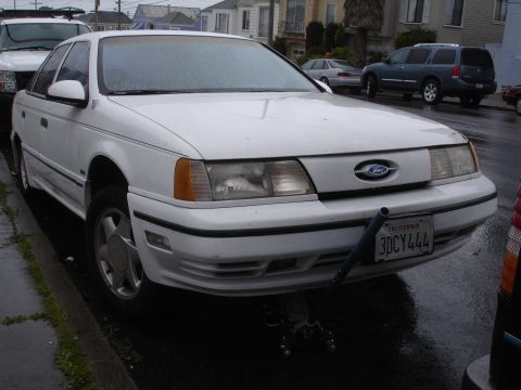 1989 Ford Taurus - A white sedan, likely a Ford Taurus from the mid-to-late 1980s, is parked on a wet street with its front end raised by a jack.
