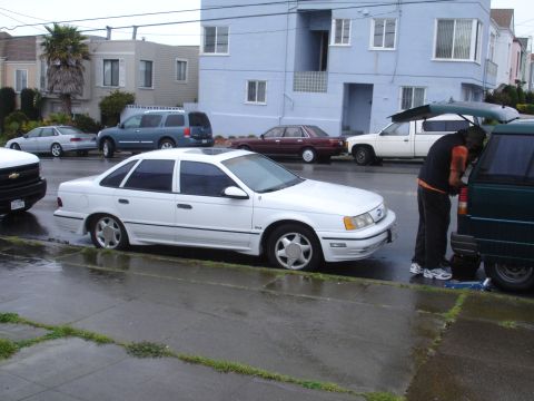 1989 Ford Taurus - A white sedan is parked on a wet street next to other vehicles and a building.