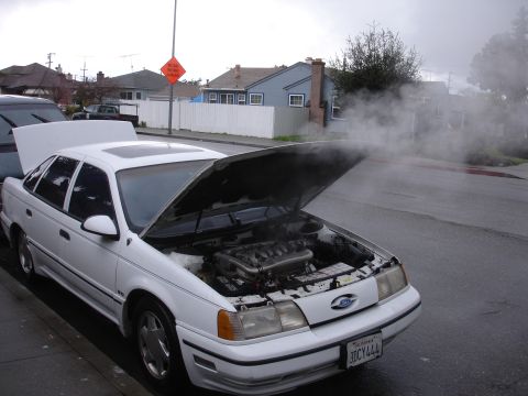1989 Ford Taurus - A white sedan is parked on the side of the road with its hood open, emitting steam.