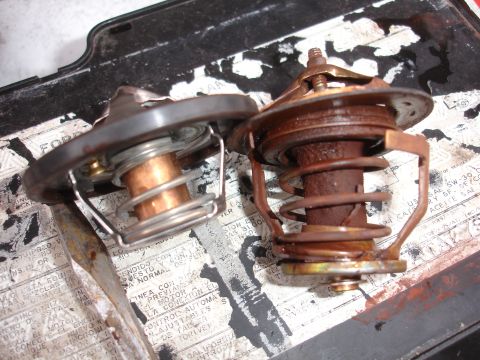 1989 Ford Taurus - Comparison of a clean thermostat and a rusted one, resting on a surface with visible text.