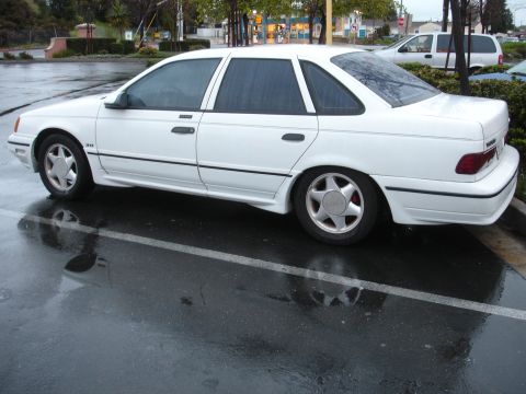 1989 Ford Taurus - A white sedan is parked in a lot on a rainy day, showing its side profile and custom wheels.