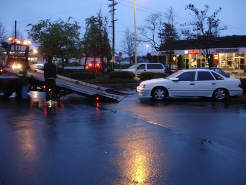 1989 Ford Taurus - A white sedan, likely from the late 80s or early 90s, is being loaded onto a tow truck on a rainy evening.