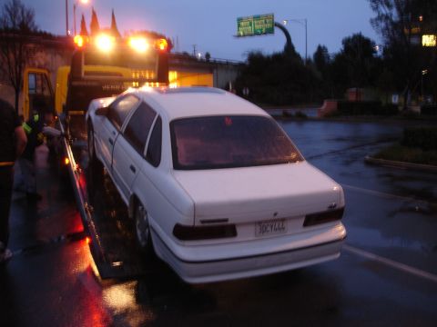 1989 Ford Taurus - A white 1989 Ford Taurus is loaded onto a tow truck as dusk settles and the road reflects the vehicle's lights.