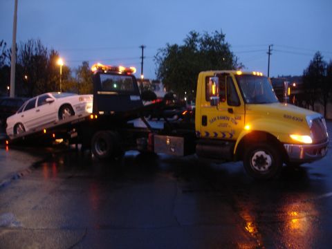 1989 Ford Taurus - A tow truck transports a white sedan on its flatbed under a twilight sky.