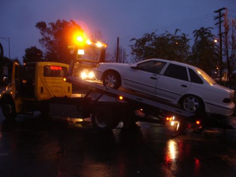 1989 Ford Taurus - A white 1989 Ford Taurus is loaded onto a tow truck on a wet evening.