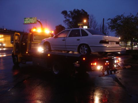 1989 Ford Taurus - A white 1989 Ford Taurus is loaded onto a tow truck at night on a wet street.