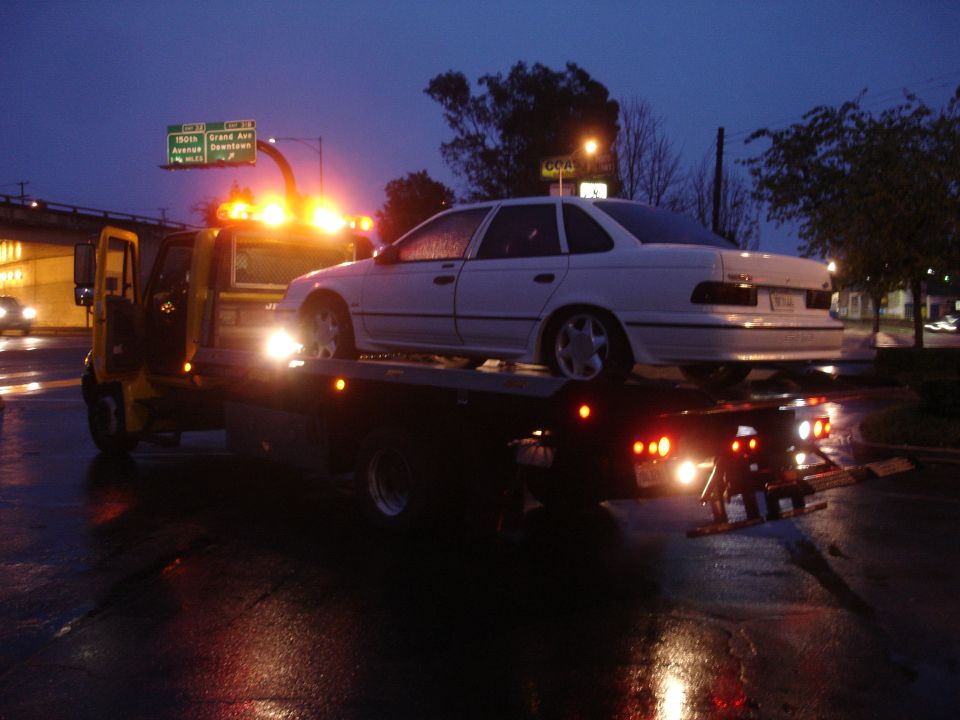 1989 Ford Taurus on a flatbed tow truck at night, wet road reflecting lights
