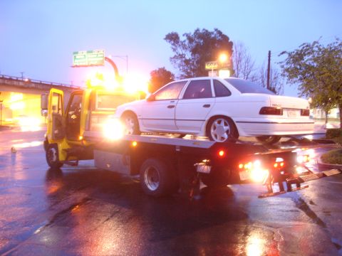 1989 Ford Taurus - A white 1989 Ford Taurus is loaded onto a tow truck on a wet road at dusk.