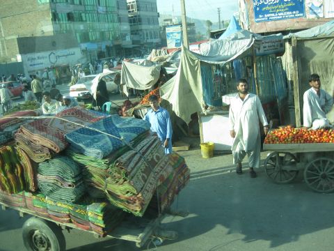 2003 Jeep Wrangler - A bustling street market scene in Afghanistan, vendors, shoppers, and various goods for sale, including carpets and produce.