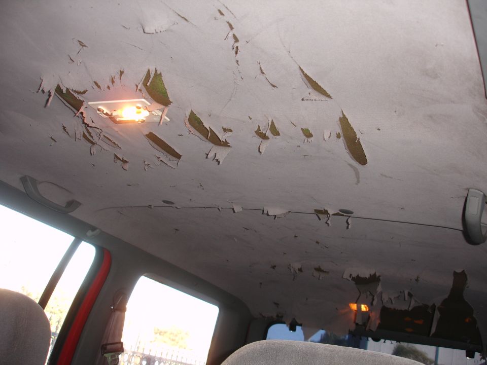 Interior of a vehicle with a damaged headliner, showing large sections of material peeling away to reveal the underlying structure. A dome light is illuminated