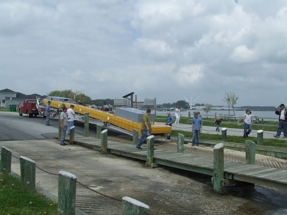 People gather around a boat ramp and dock, with a trailer and a red truck visible in the background